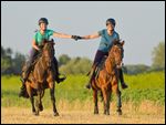 Zwei Reiterinnen auf Reitponys galoppieren Hand in Hand über ein Stoppelfeld 