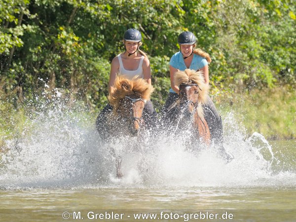 Zwei junge Reiternnen auf IslÃ¤ndern galoppieren in der Isar sÃ¼dlich von MÃ¼nchen